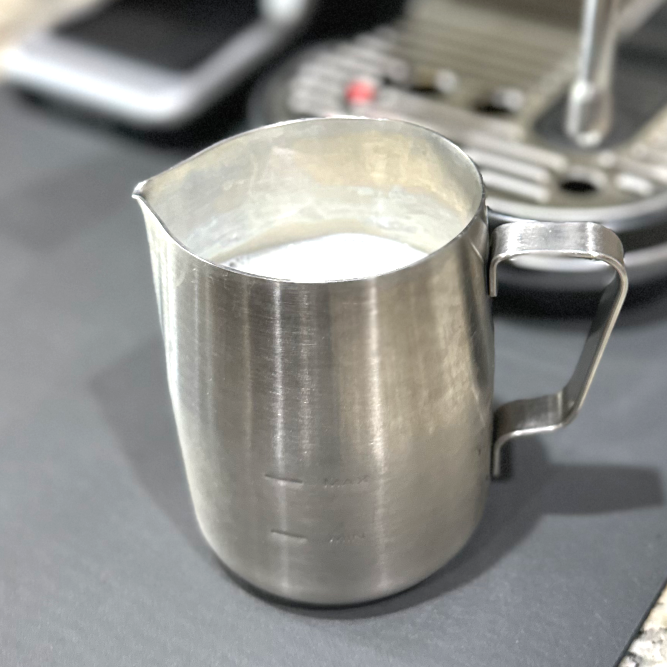 Stainless steel milk pitcher filled with milk beside a Breville Bambino Plus espresso machine and steam wand