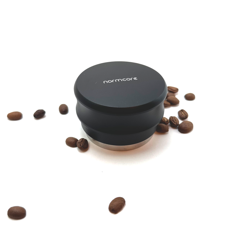 Normcore spring‑loaded palm tamper surrounded by coffee beans, designed to apply level, consistent tamping pressure.